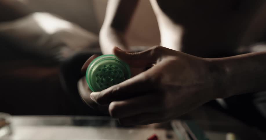 Man puts pieces of a substance in a round green plastic grinder then twists the grinder to grind it