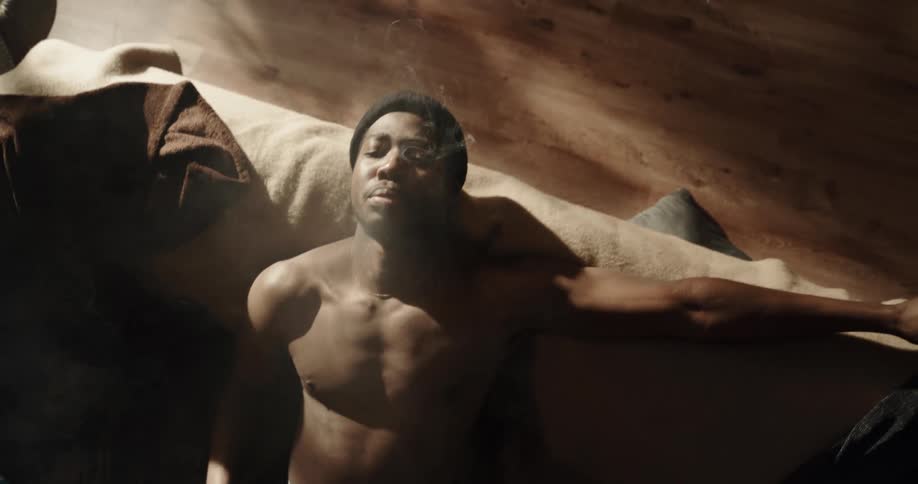 Shirtless black man sitting on the couch wearing a beanie and smoking a joint