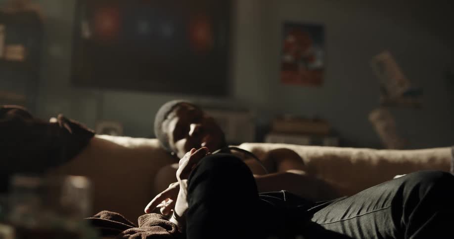 A teenage boy sits on a couch and shoots drugs in to his arm and then flops his head back