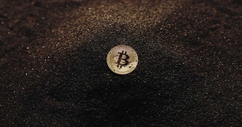 Close up of the shiny engraved surface of golden bitcoin physical coins over dark sand blowing in the wind