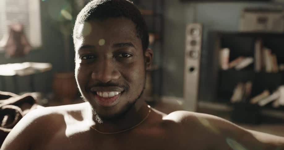 A shirtless man raises his head to look into the camera and smiles for a moment before lowering his gaze