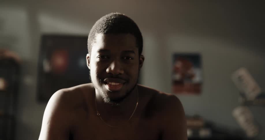 A shirtless man sitting in a house smiles while gazing into the camera