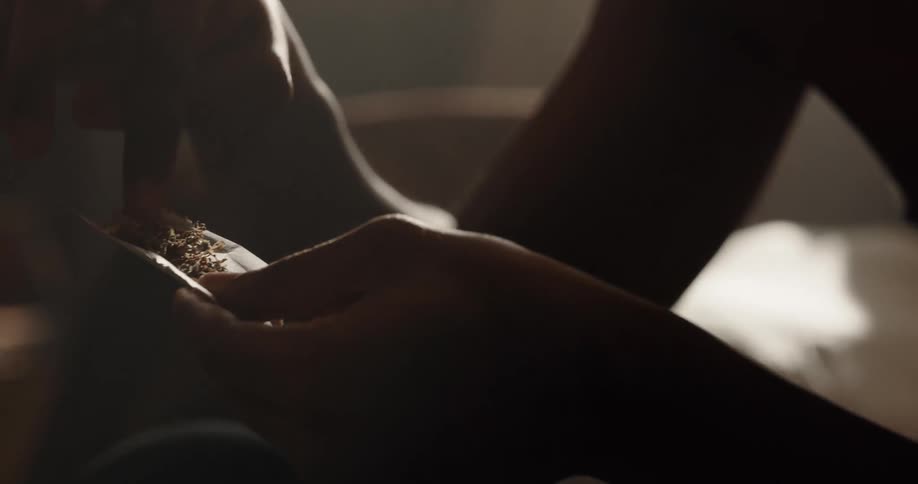 Shirtless black man sitting on the couch pouring cannabis into a joint