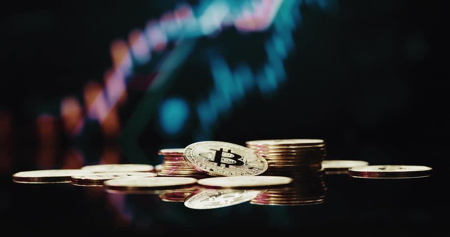 Pile of physical engraved golden coins with bitcoin logo in front of a screen showing a graph