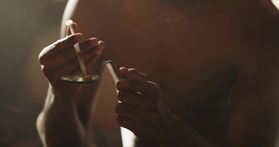 Man sitting on a couch at home uses a syringe to fill a bent spoon to cook drugs