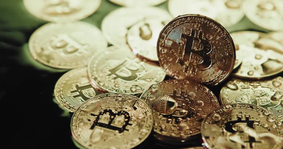 A number of shiny coins with the bitcoin logo on them are piled on a very wet surface