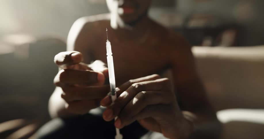 Black man sitting on the couch at home holding a drug syringe and preparing it to inject
