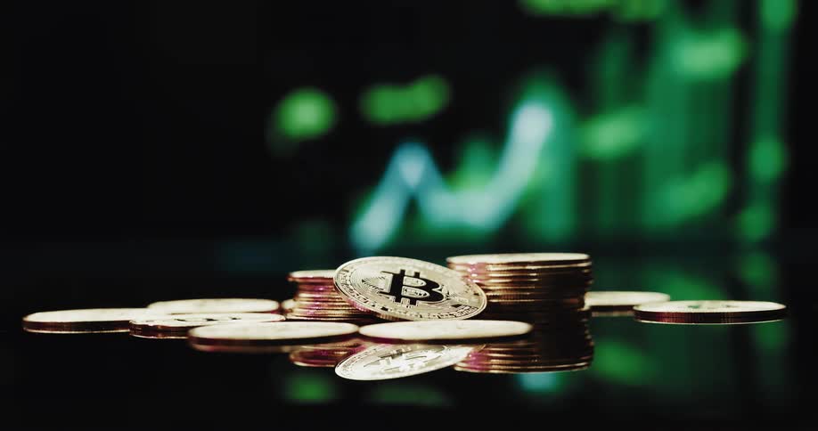 Gold plated bitcoins on a reflective surface with a computer screen displaying a climbing graph