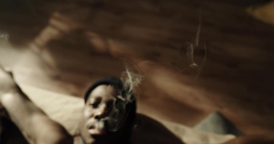 Young man laying in bed at home smokes a cigarette watching the smoke rise at morning