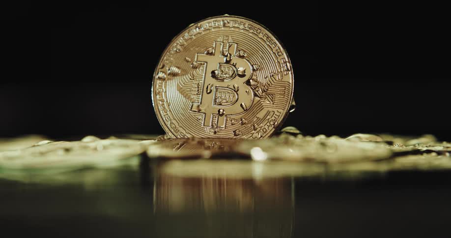 A gold bitcoin is standing on edge above moving water with water droplets on it
