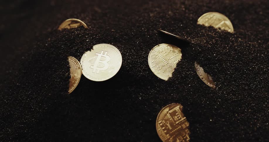 A number of shiny coins with the bitcoin logo on them are partially buried in black powder being blown around by wind