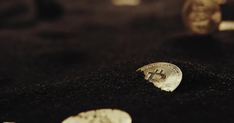 Close up of the shiny engraved surface of golden bitcoin physical coins over dark sand blowing in the wind