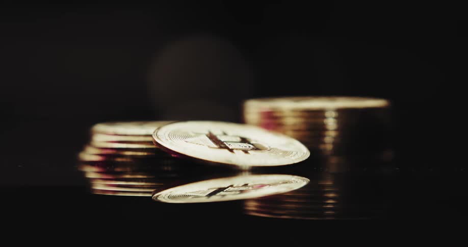 Stacked gold bitcoins are on a mirror and a single bitcoin is tilted in front of the stacks