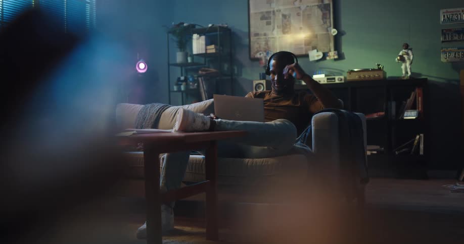 Black man sitting on the couch in his living room with a laptop on his lap putting on a headset and talking to someone on a video chat