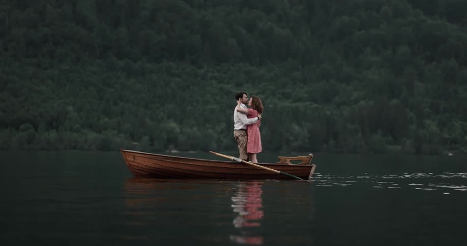 A man and woman kiss while standing in a small boat on a lake during the day