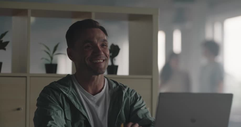 A man sits at home at a table in front of a computer and sighs as he puts papers away and then talks and laughs with someone as his teenagers walk behind him