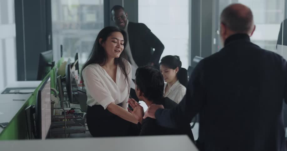 Businesswoman with white shirt and black skirt sits on desk talking with businessman wearing black blazer jacket in the office