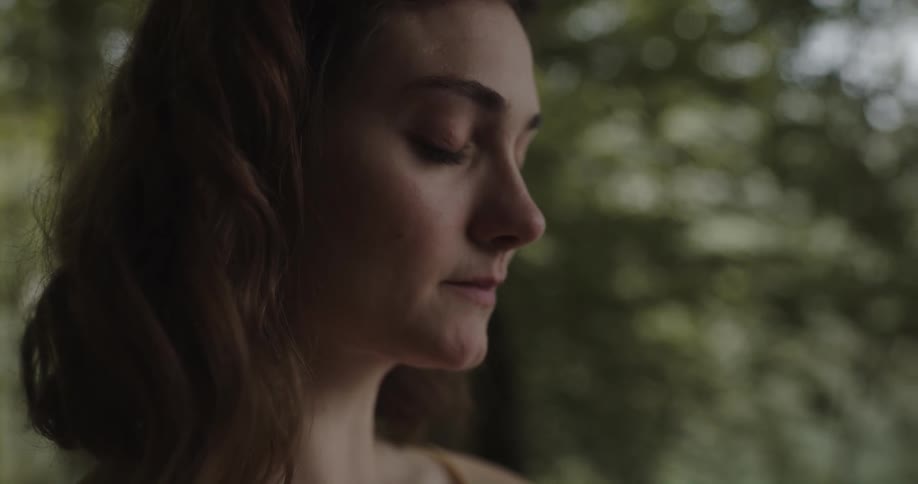 A woman standing amids to grove of trees looks out across the distance with a solemn expression on her face