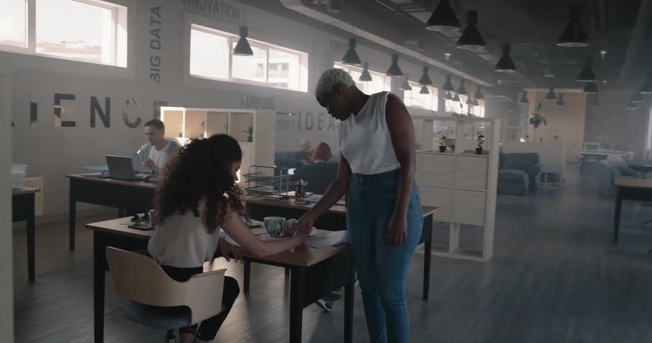 Woman walks past people sitting at desks and gives a man encouragement and stops to talk to a woman