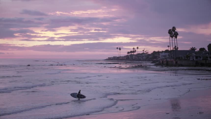 A person is exiting the water of a beach carrying a surfboard at sunset