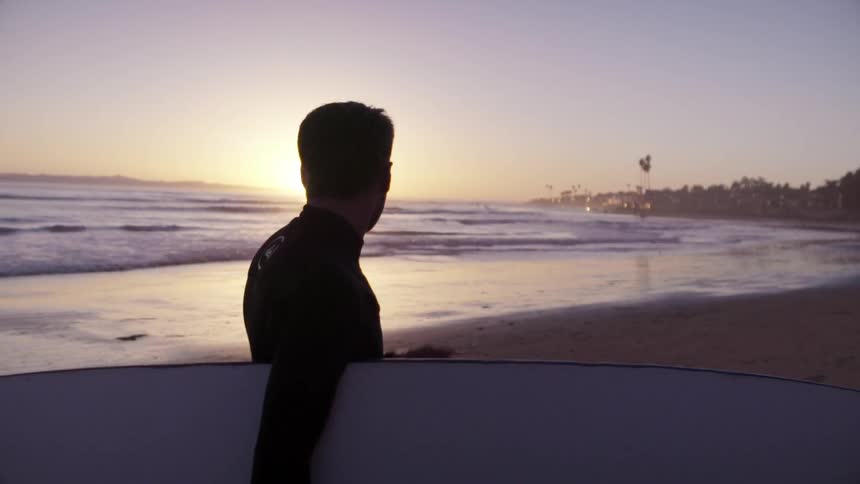 A man holding a surfboard looks over at the sunset on the beach