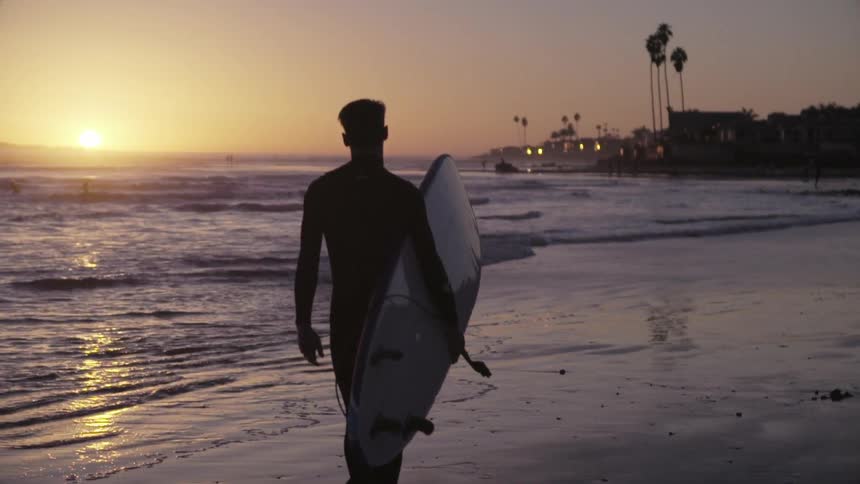 Person carrying surfboard walks on the beach at sunset
