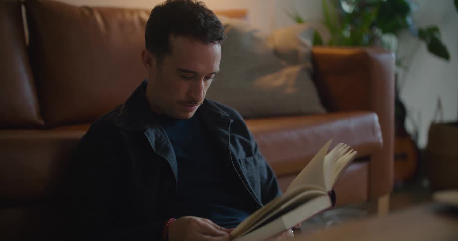 A man sitting on the floor in front of his couch turns the page of a book he is reading