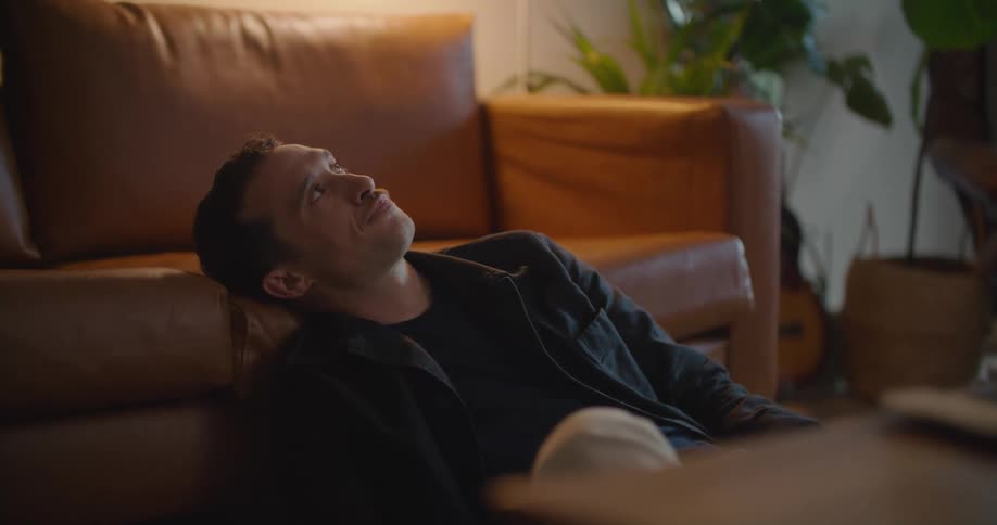 Thoughtful white man sitting on the floor leaning his head on the couch and using his smartphone