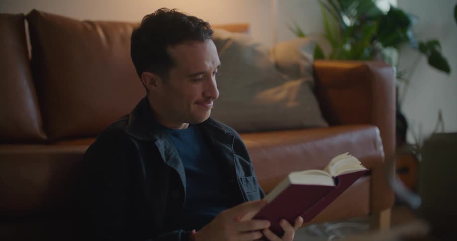 Man with naval blue jacket and t shirt sits on the floor and reads book leaning against the brown couch in the living room