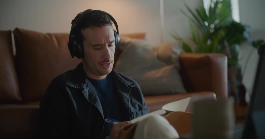 White man sitting on the floor in his living room wearing a headphone and talking to someone while holding a book