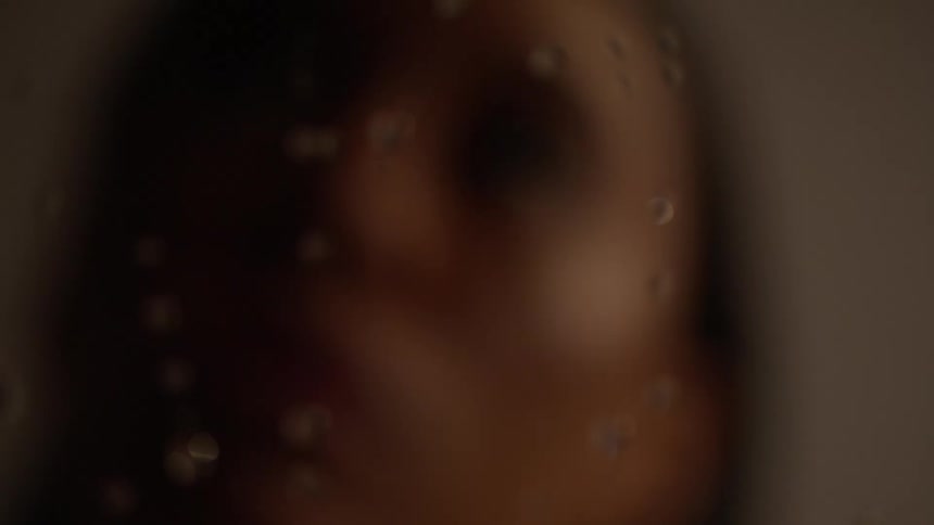 A woman looks upwards while water droplets roll down a piece of glass in front of her