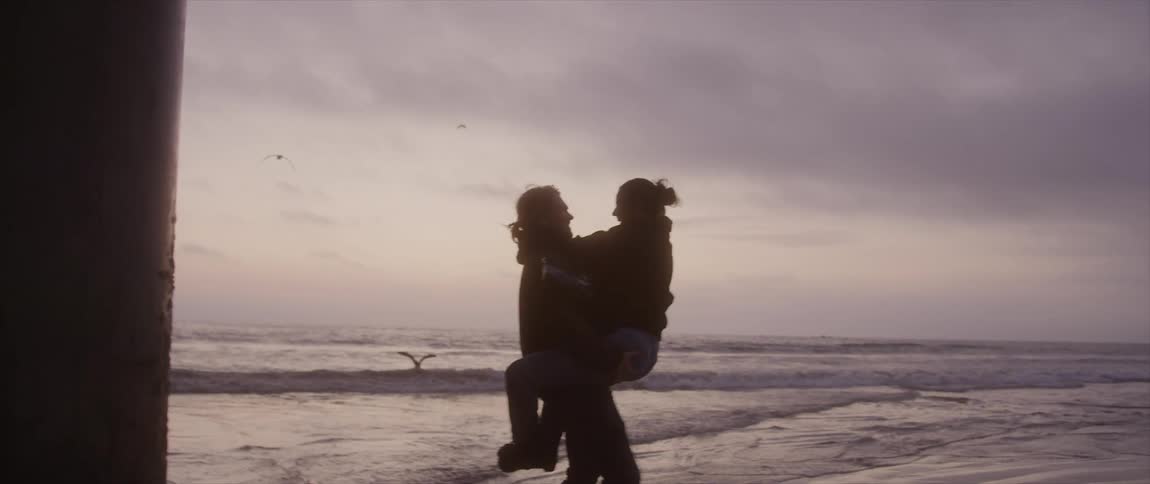 Man with long hair carrying woman in arms and turning around is silhouetted against the purple sky next to pier on the beach at dusk