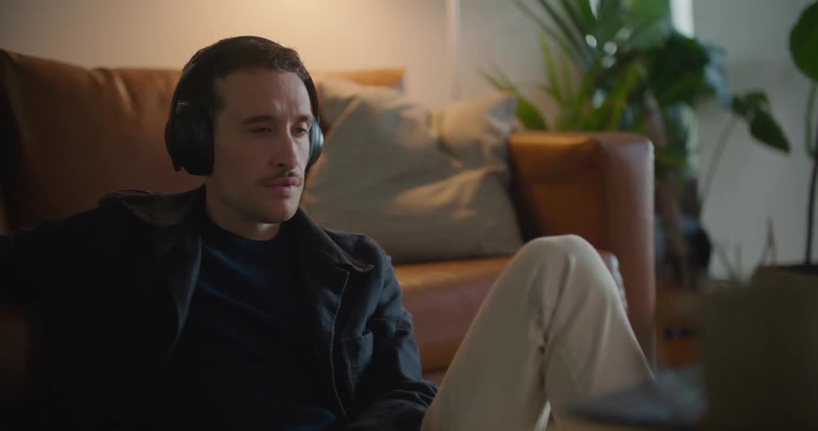 Man with naval blue jacket shirt brown pants and headphones watches tablet sitting on floor leaning against brown couch in living room