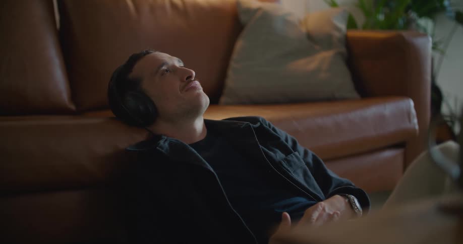 Young man reclined on a couch sitting on the living room floor listening to music on hearphones