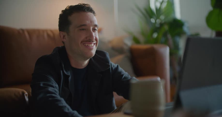 Man with mustache naval blue jacket and shirt sits on floor leaning against brown couch and making a video call on tablet