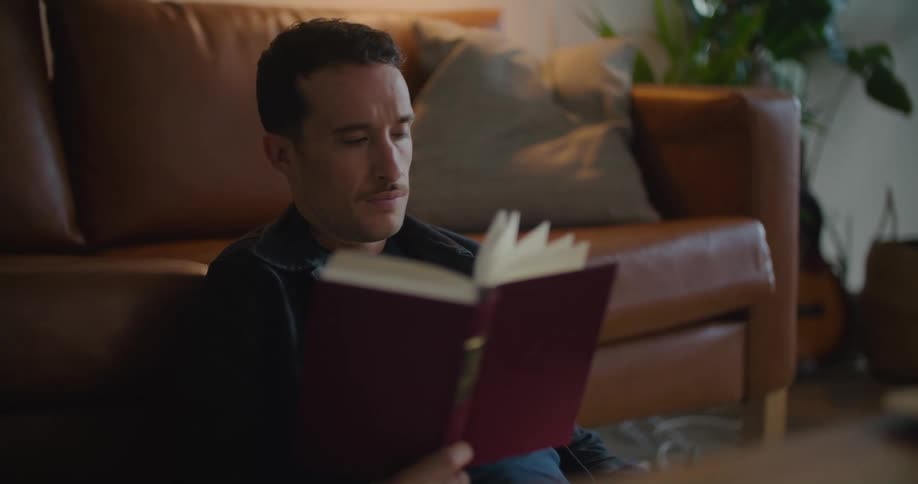 Man sitting on the living room floor reading a book while checking his phone