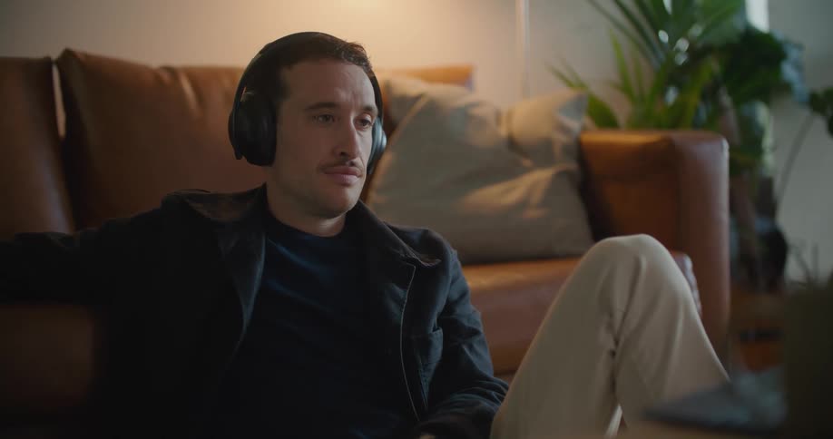 A man sits on the floor in front of his couch with headphones on and watches his laptop while swiping on his cell phone