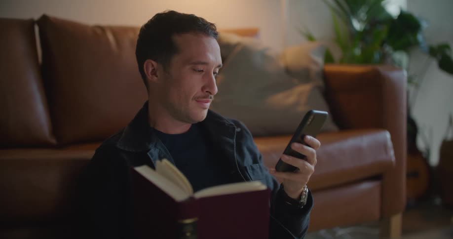 A man sits on the floor in front of his couch with a book while texting on his phone