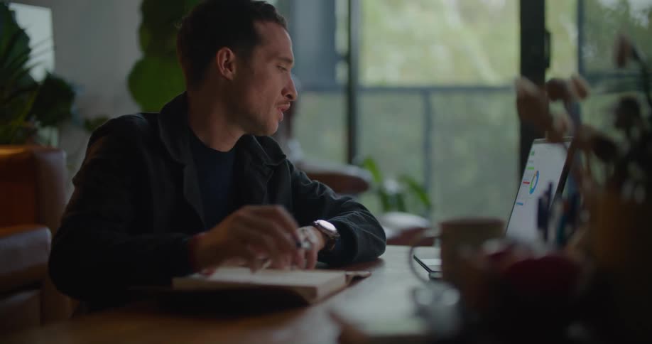 A man is sitting at a table in front of a laptop computer and writing in a notebook