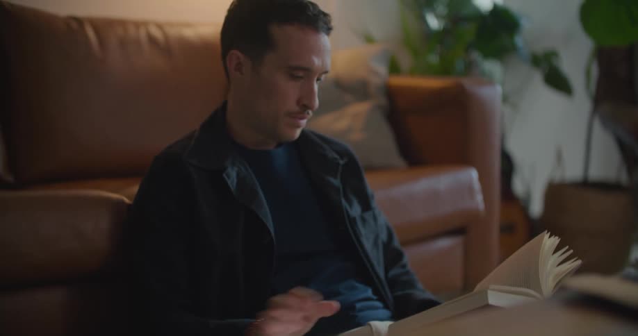 Man with naval blue jacket shirt and brown pants uses smartphone and reads book sitting on floor leaning against brown couch