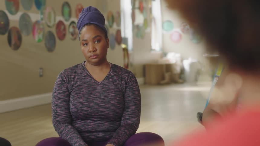 A woman wearing a wrap on her hair sits on the floor while listening to another woman