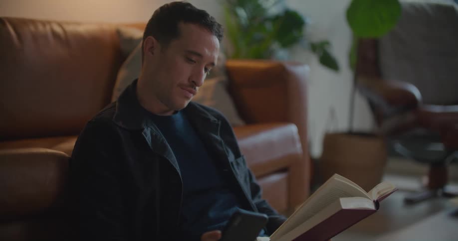 Man sitting on the floor leaning on the couch behind him reading a book and using his smartphone