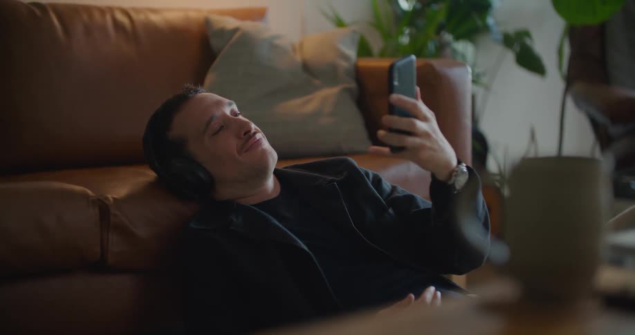 Young man reclined on a couch sitting on the living room floor listening to music on headphones