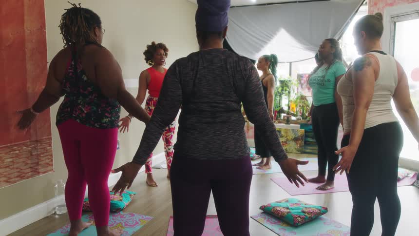A group of women stand in a circle and stretch their arms out to their sides as they listen to an instructor