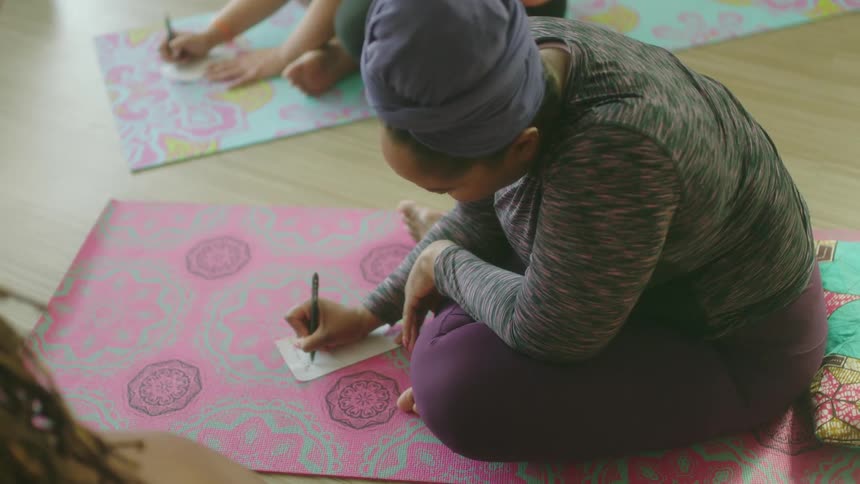 A group of women write on small sheets of paper while sitting on yoga mats on the floor