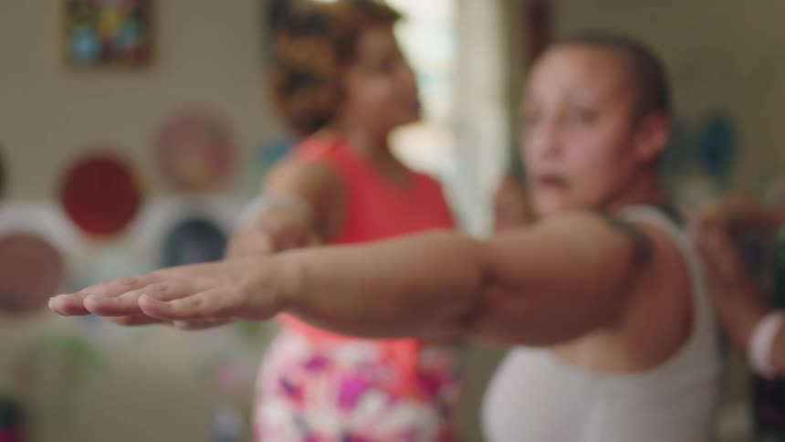 A woman stands holding a yoga position as another woman adjusts her arm and another woman talks to them