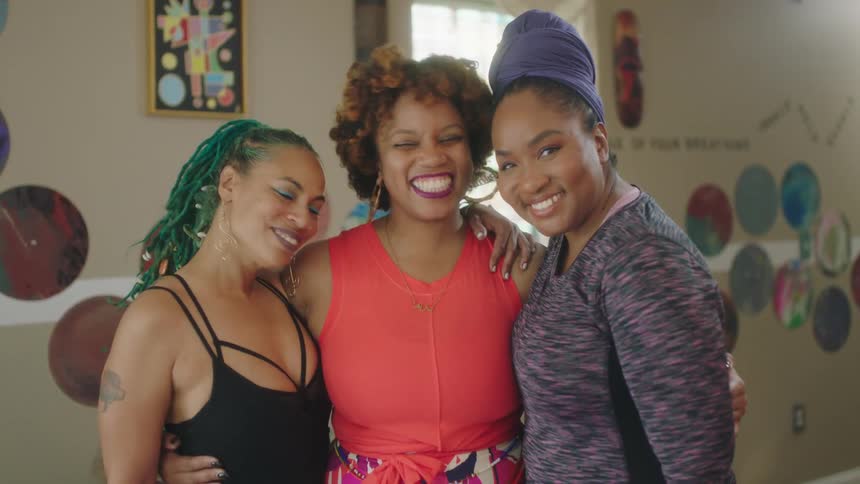 Three women stand close together with their arms around each other and smile