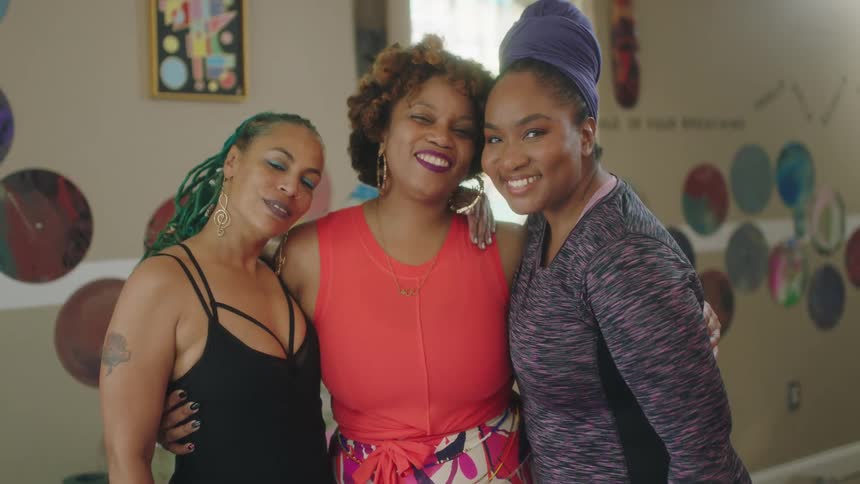 Three women stand in a room together and put their arms around one another and smile