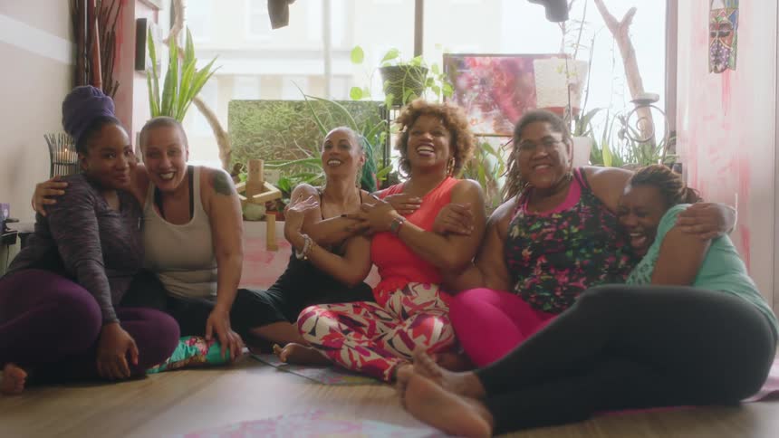 Group of barefoot women wearing sports clothes sitting over mats in the floor hugging and laughing