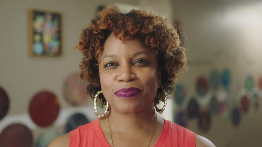 Black woman with curly hair wearing makeup and earrings standing indoors and smiling